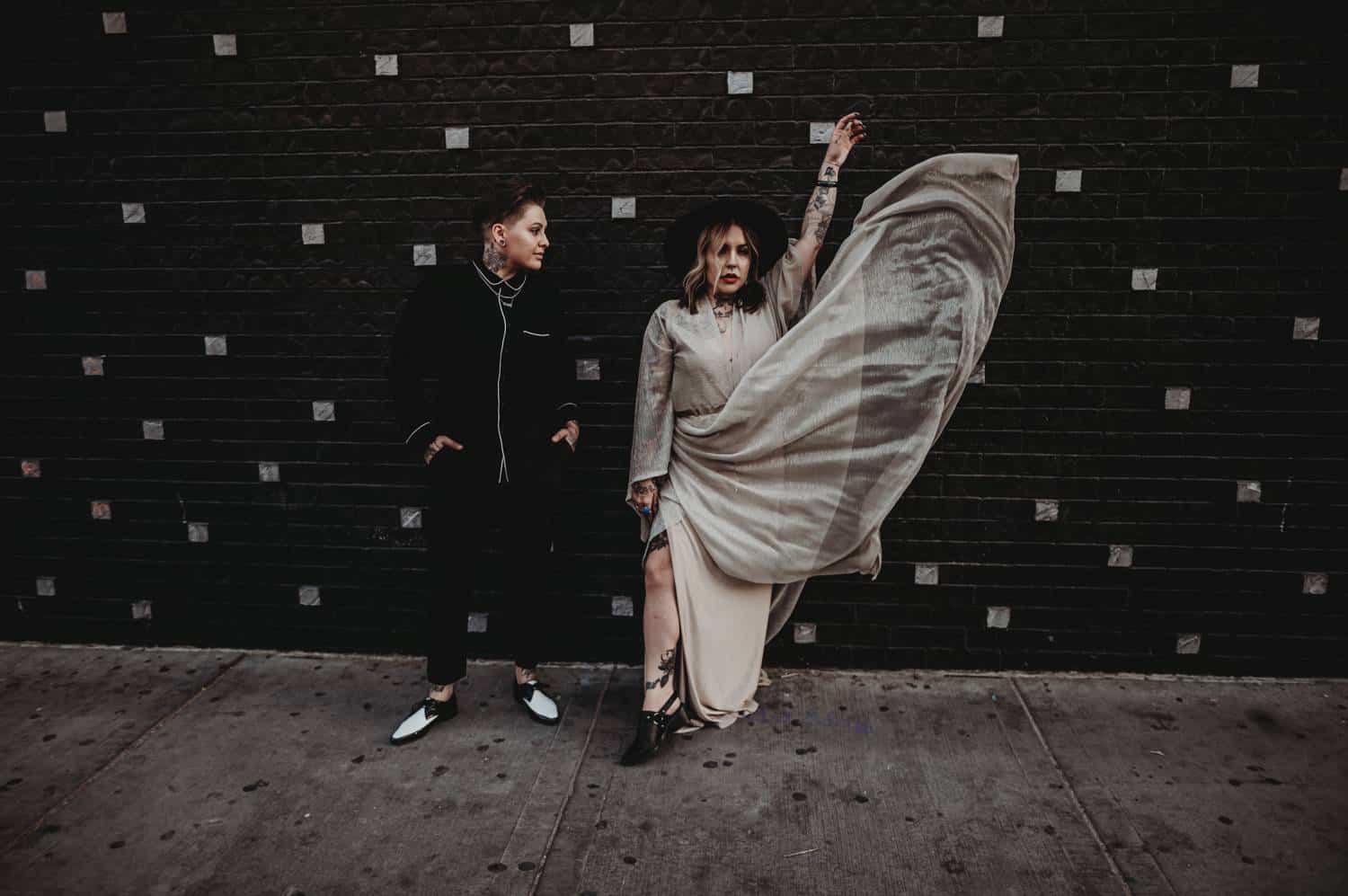 A couple stands side-by-side against a black metal wall. The person on the left is wearing a modern black suit. The person on the right is wearing a sparkly, flowing dress, which is being tossed in the air.