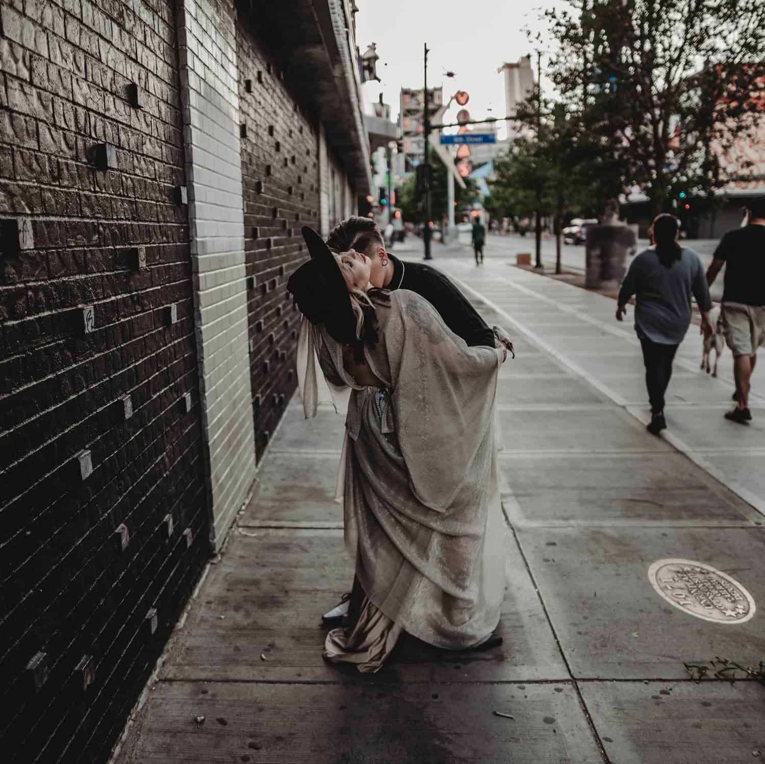 A couple stands in the middle of a Las Vegas sidewalk as tourists wander by. One partner is wearing black and dipping the other partner, who wears a floppy hat and a shimmery white gown.