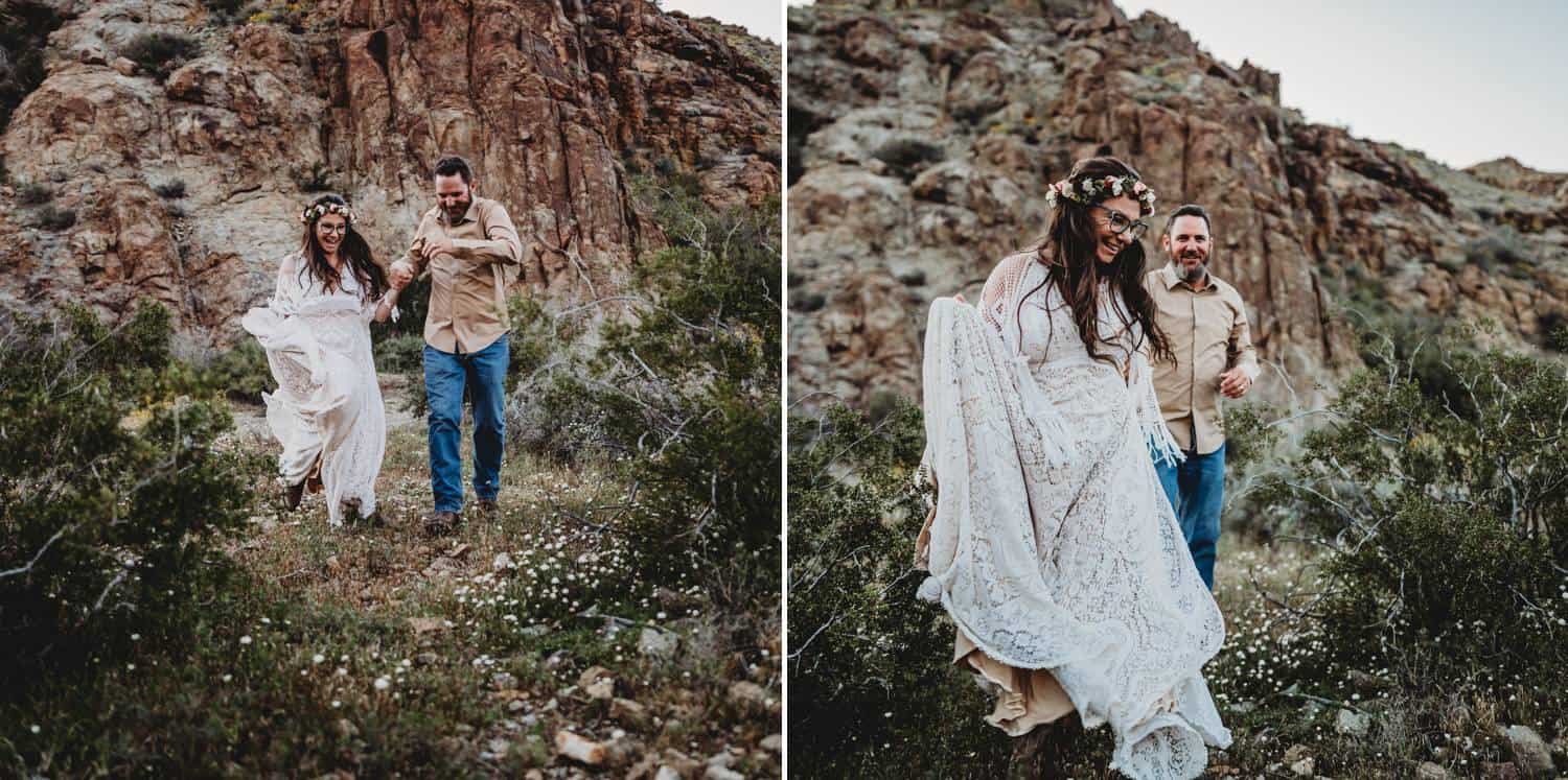 A white-gowned bride holds hands with her more casually dressed groom as they skip together through the grass at the foot of red desert mountains.