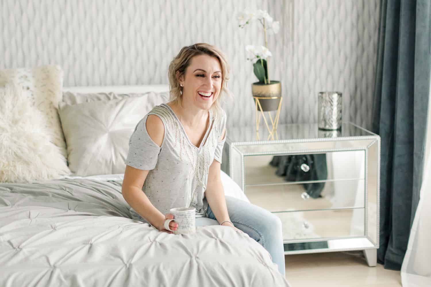 A woman holding a coffee mug sits on a bed laughing in a bright white bedroom