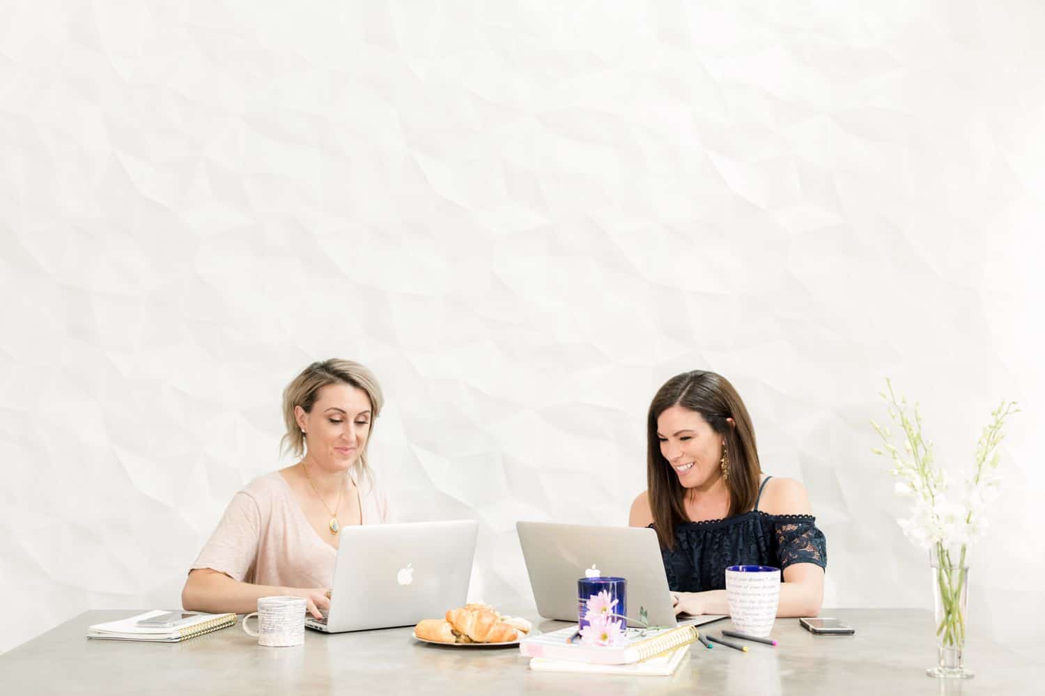 Two business women work at a large desk in a white room.