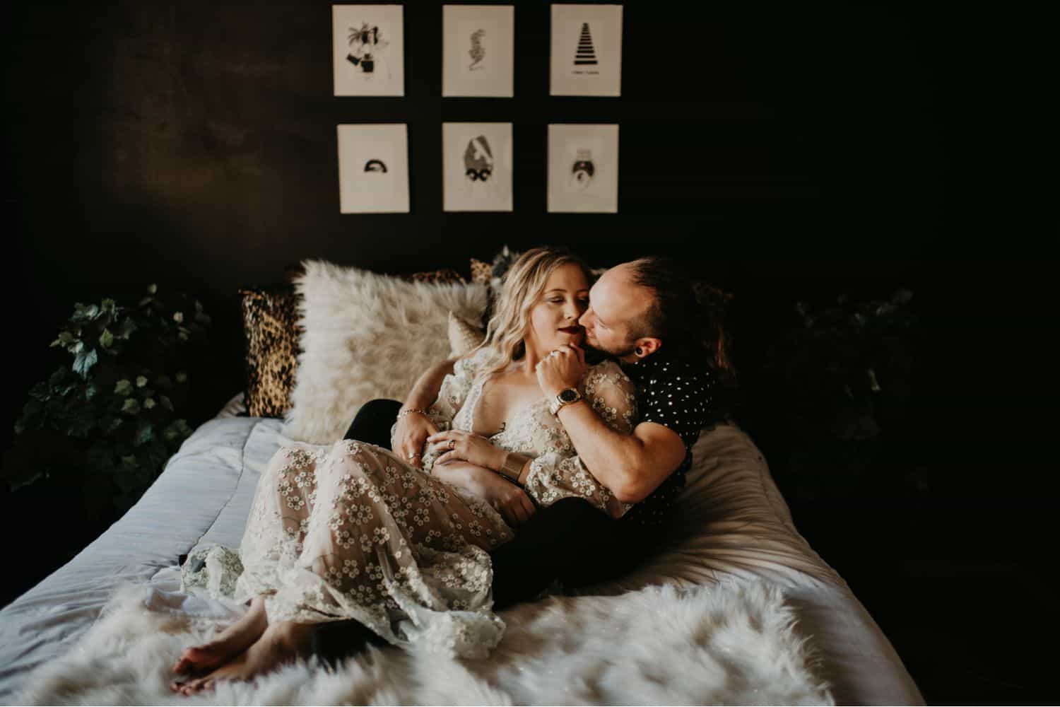 A woman in a white lace dress reclines on a bed in the arms of a man in a black shirt. Learn how to take low light photos like Shelby Laine Photography.