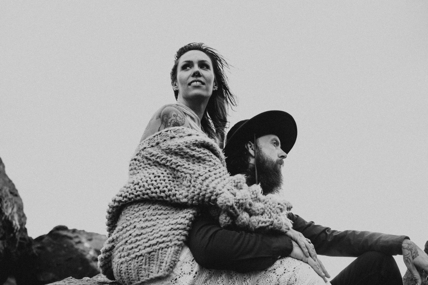 A woman in an oversized sweater sits behind a man with a beard as they sit outside against a gray sky.
