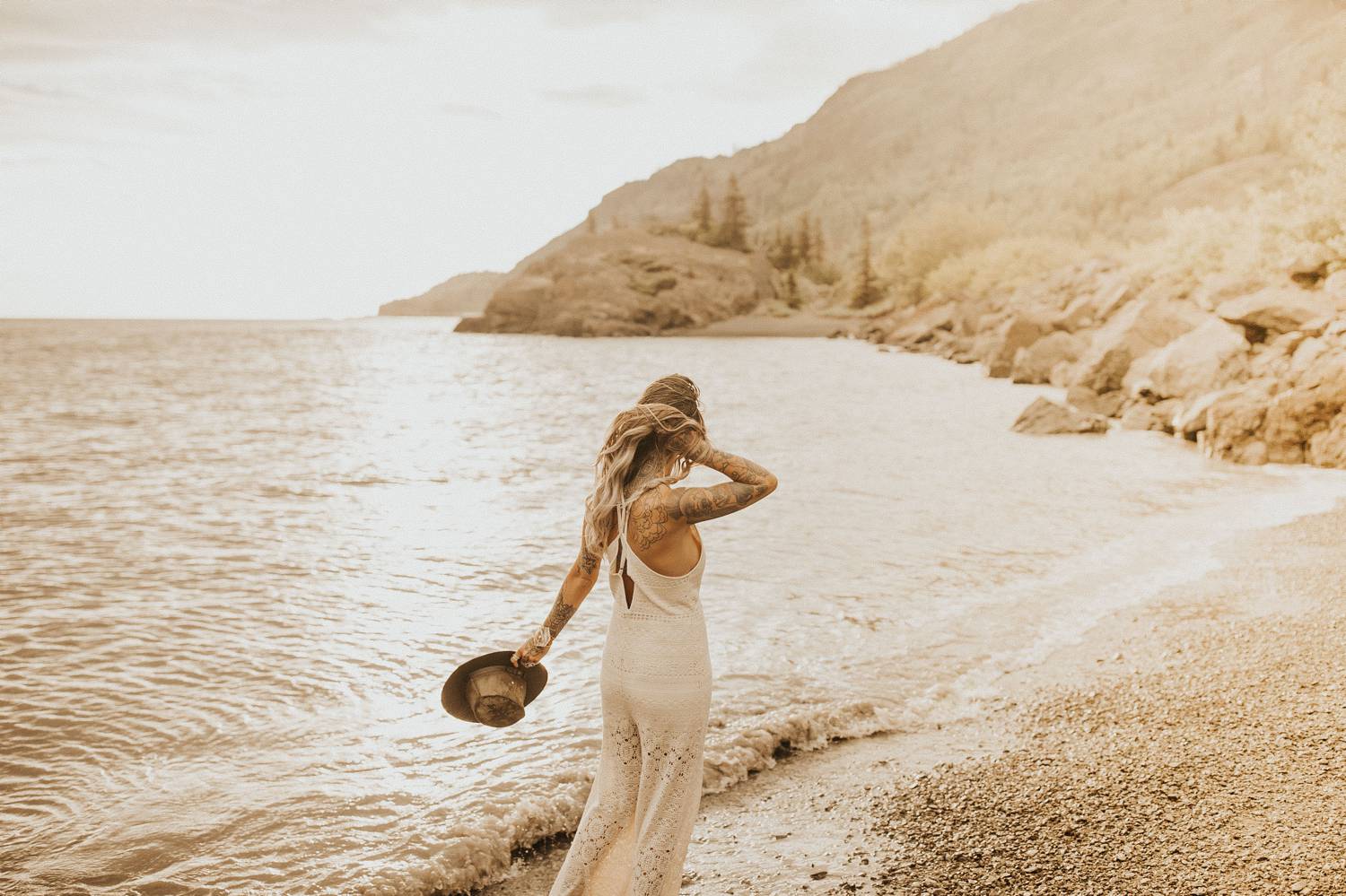 A woman in a white lace jumpsuit carries a felt had in one hand while holding her hair back with the other. She is strolling along the shore of a mountain lake.