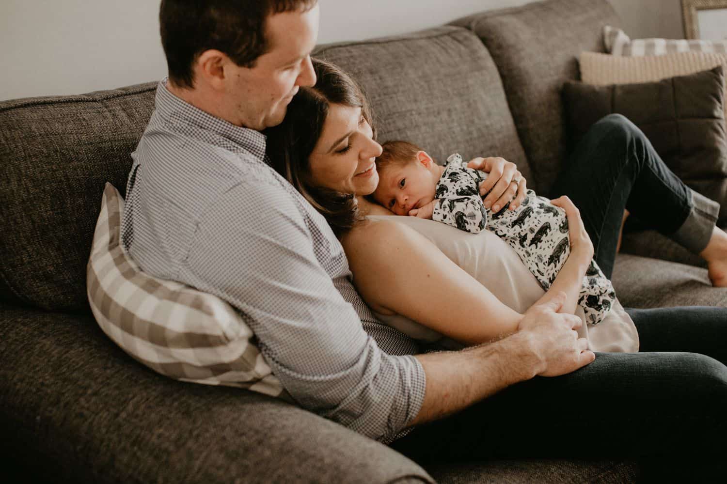 Lifestyle Newborn Photos: Dad, Mom, and Baby sit on a linen sofa in a sunlit room. They are snuggled one after the other, and the mom is smiling at her newborn.