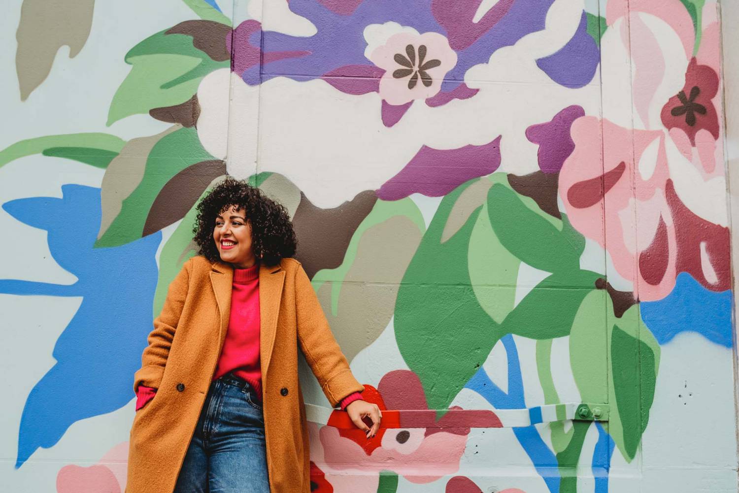 A woman business owner wearing jeans, a pink sweater, and a mustard yellow coat stands smiling against a massive wall of graffiti in Atlanta, Georgia.