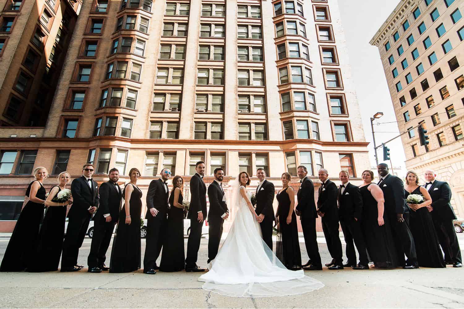 A bride and groom stand shoulder-to-shoulder with their wedding party in the middle of a city.