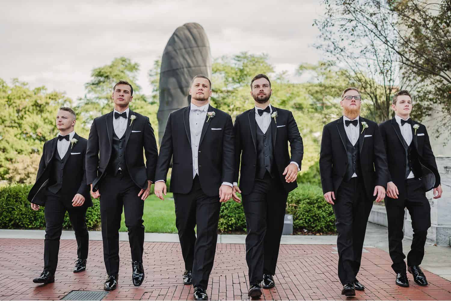 A row of groomsmen in black suits walk across a brick courtyard.