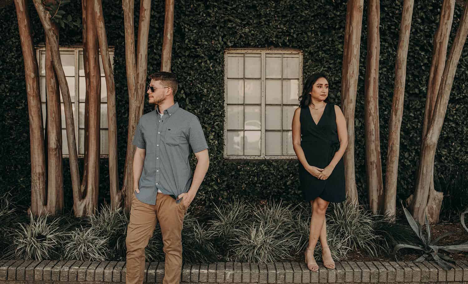 A tall man stands in front of a shady building looking to his right while a petite woman stands on a ledge above him looking to her left