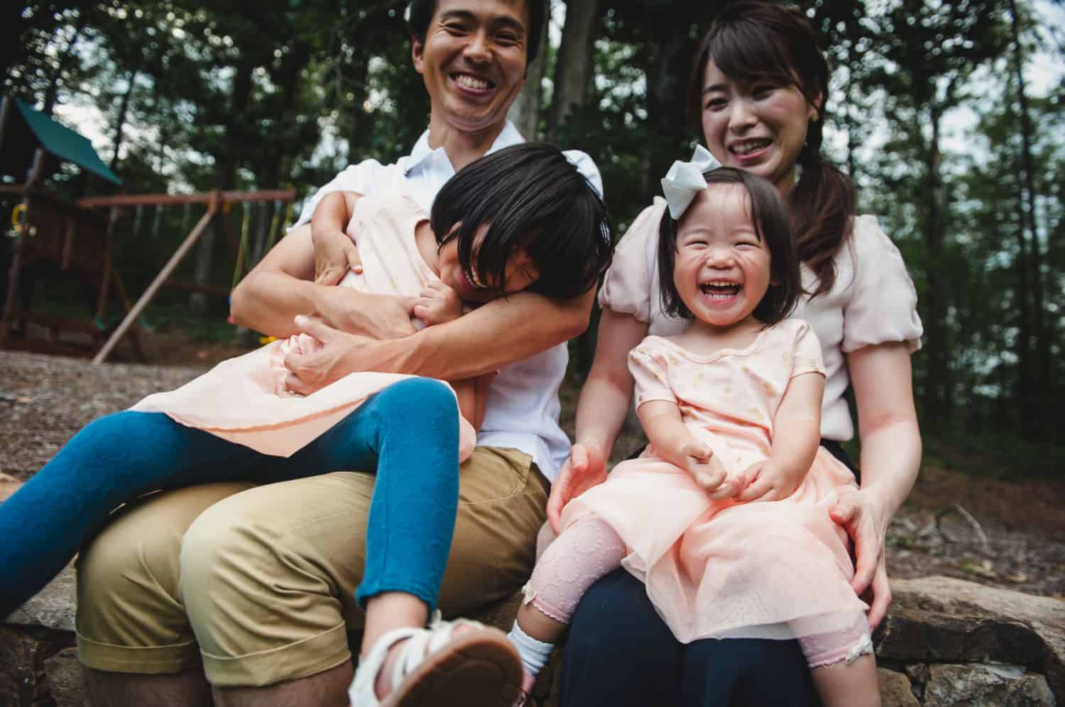 Two parents tickle their daughters while sitting together in a park.