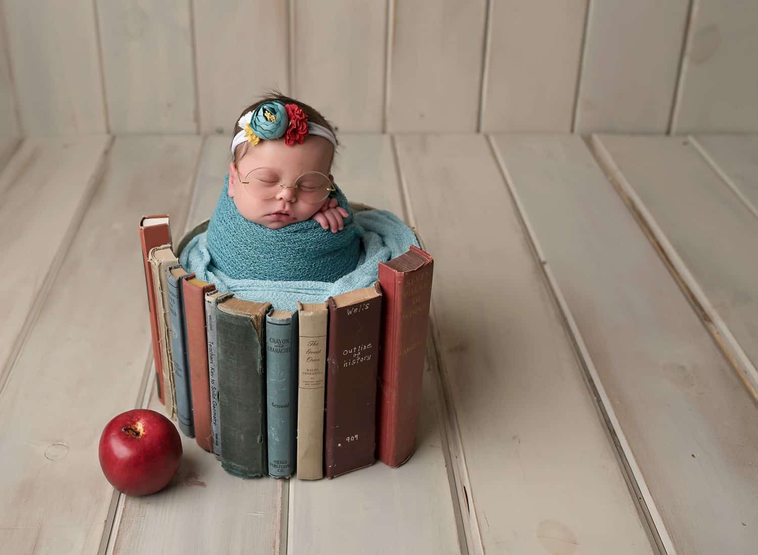 If you use only the best newborn photography props, you'll love this faux series of vintage book spines. In this photo, the "books" wrap around a newborn baby who is posed in a blue swaddle. An apple sits on the faux maple floor beneath her.