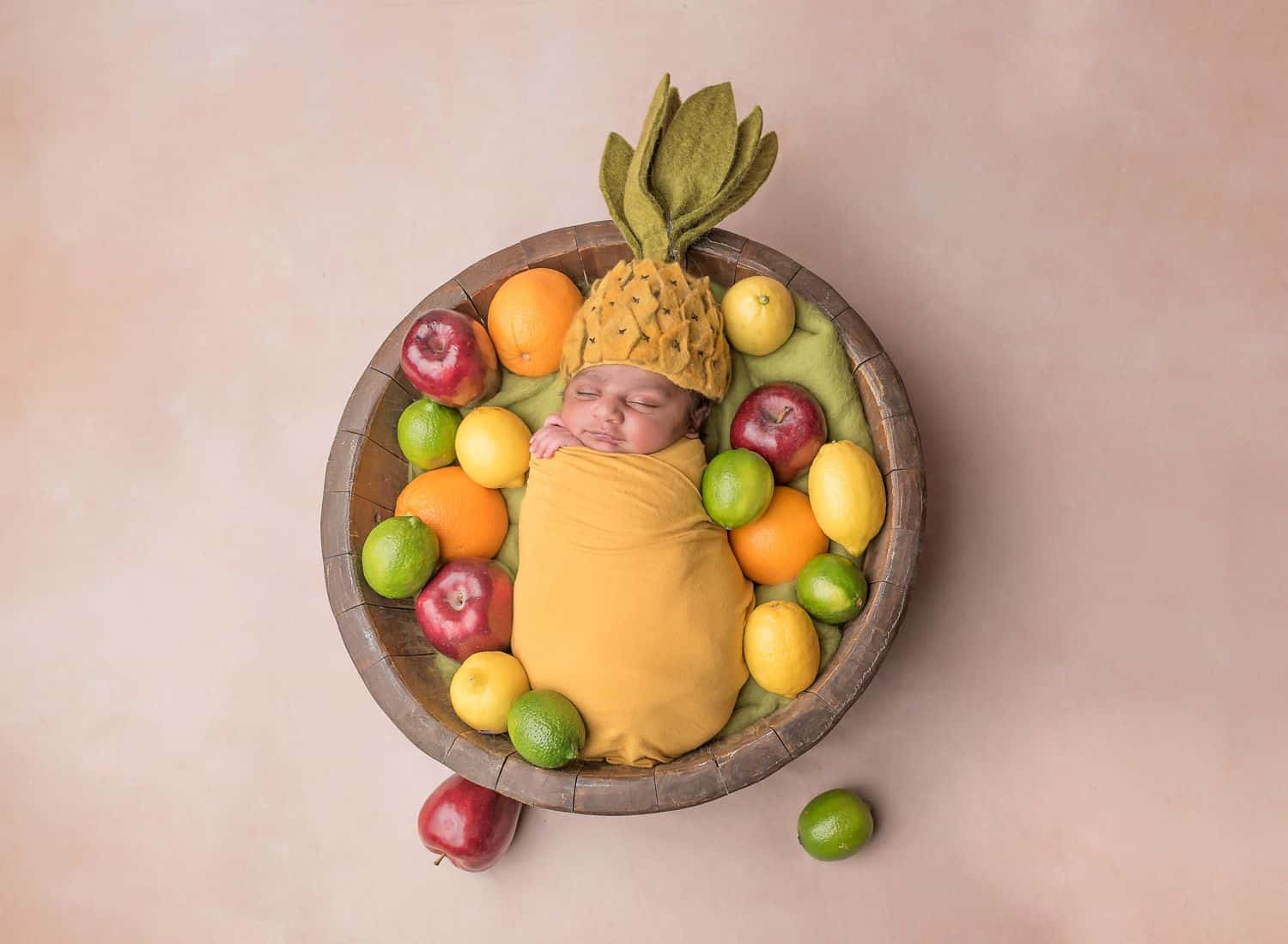 The best newborn photography props include fun themes like this fruit bowl concept. A newborn baby is wrapped in yellow and swaddled in a wooden bowl. The baby wears a felt pineapple hat and is surrounded by lemons, limes, oranges, and apples.