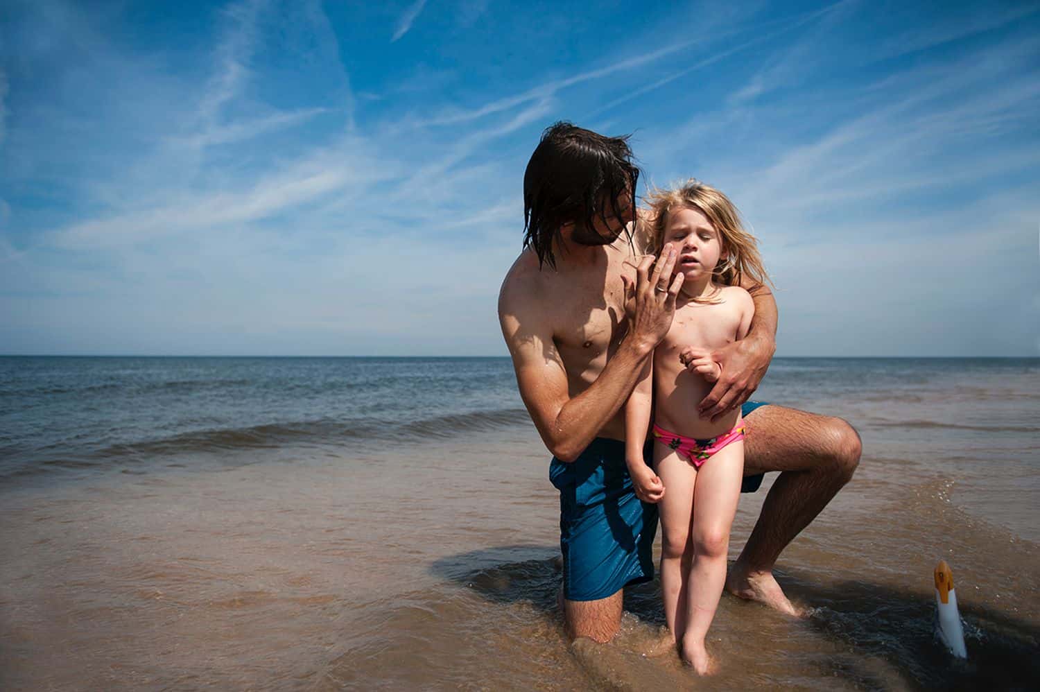 A dad wipes sunscreen on his little girl's face at the lake water's edge