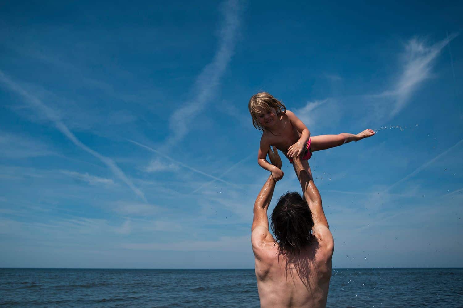 A shirtless dad tosses his young child into the blue sky above a lake