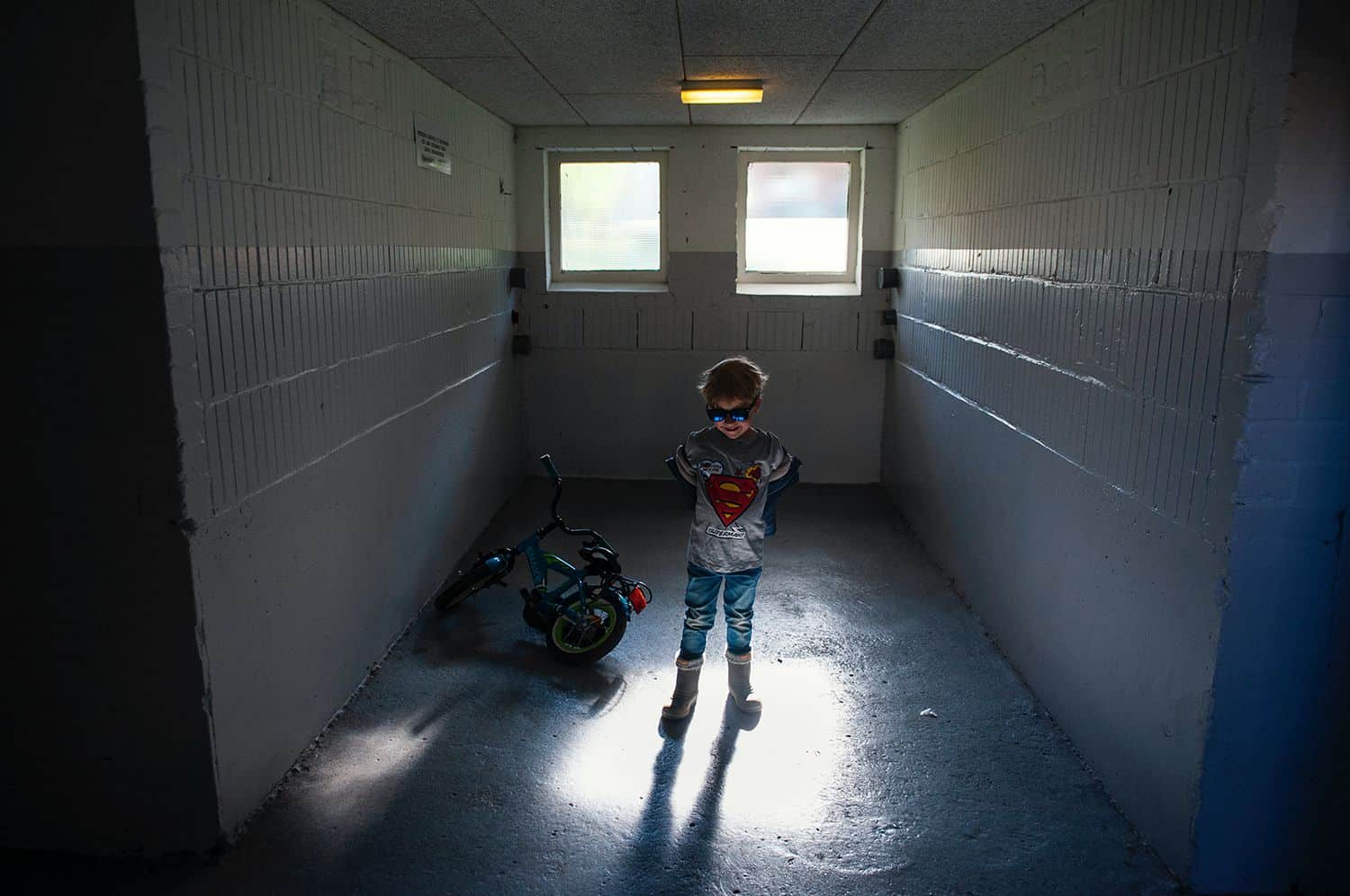 A little boy in a Superman shirt and sunglasses stands in a dim room with his hands on his hips