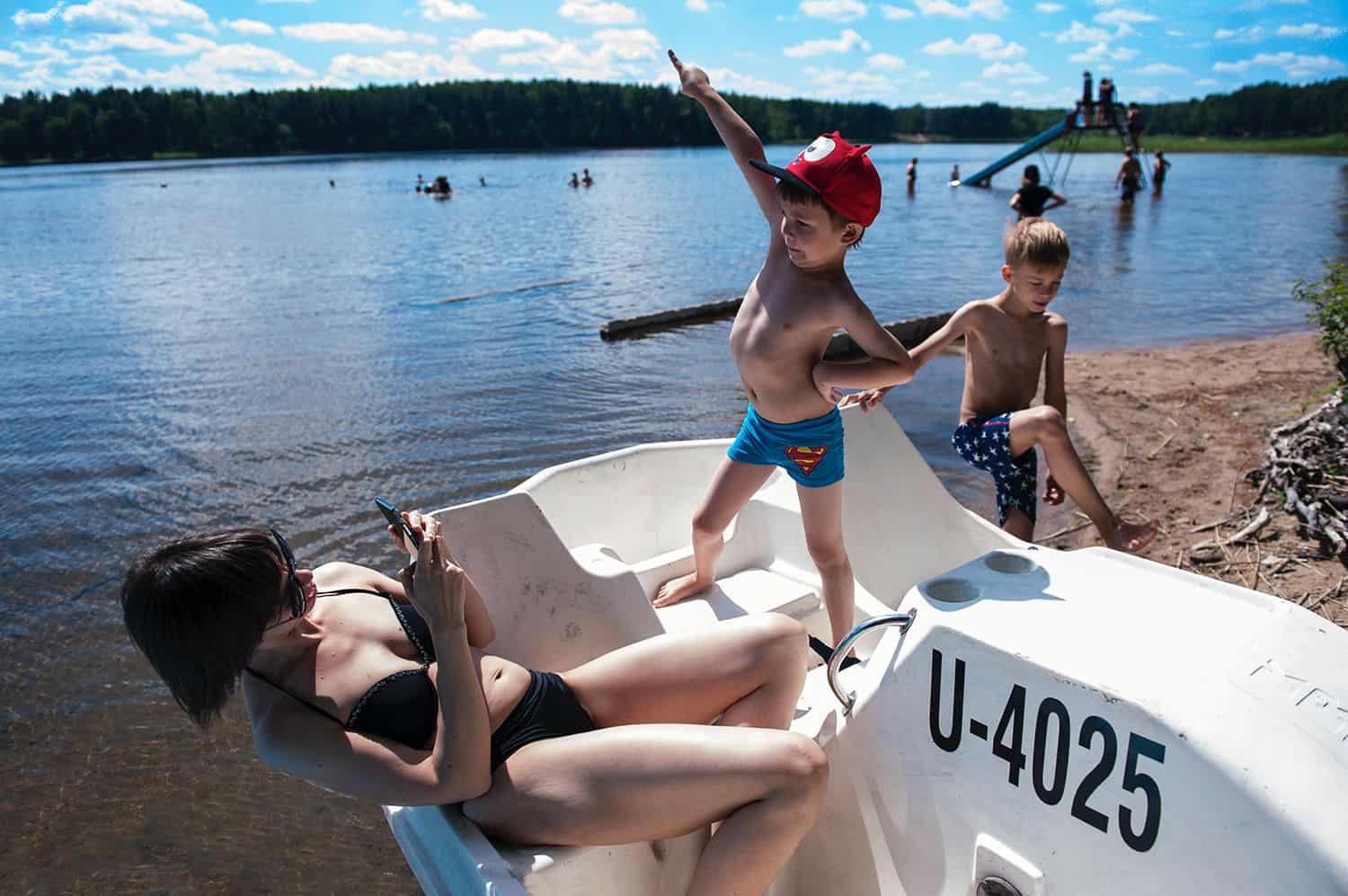 A little boy in a Superman bathing suit poses for his mom while standing on a peddle boat at the lake