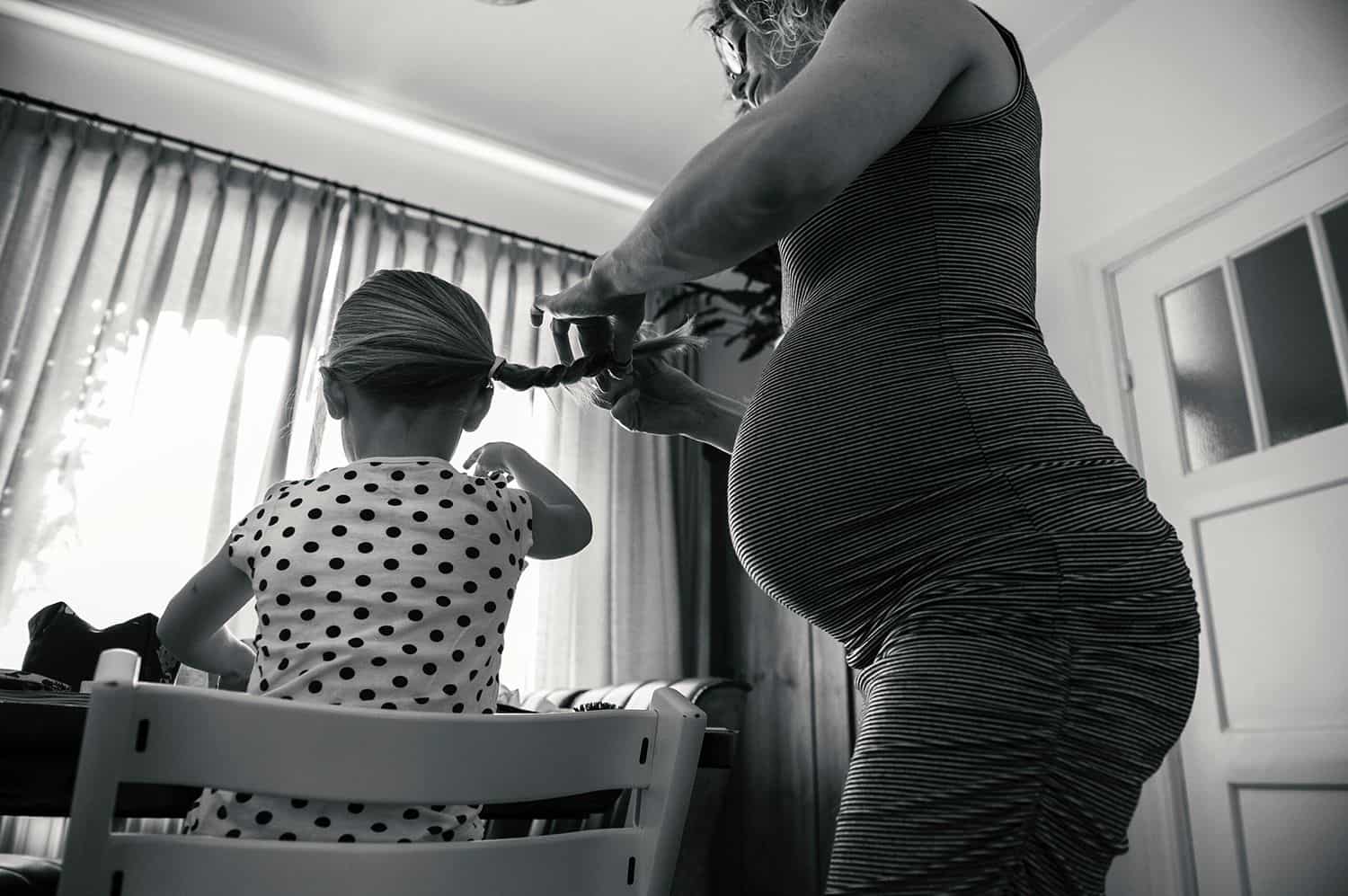 A pregnant mom braids her daughter's hair as the little girl eats breakfast