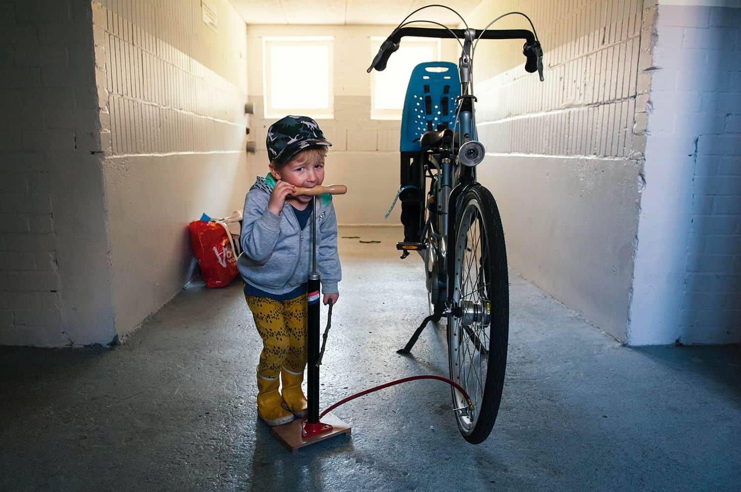 A little boy rests his mouth on the handle of the air pump he's using to inflate his bicycle tire