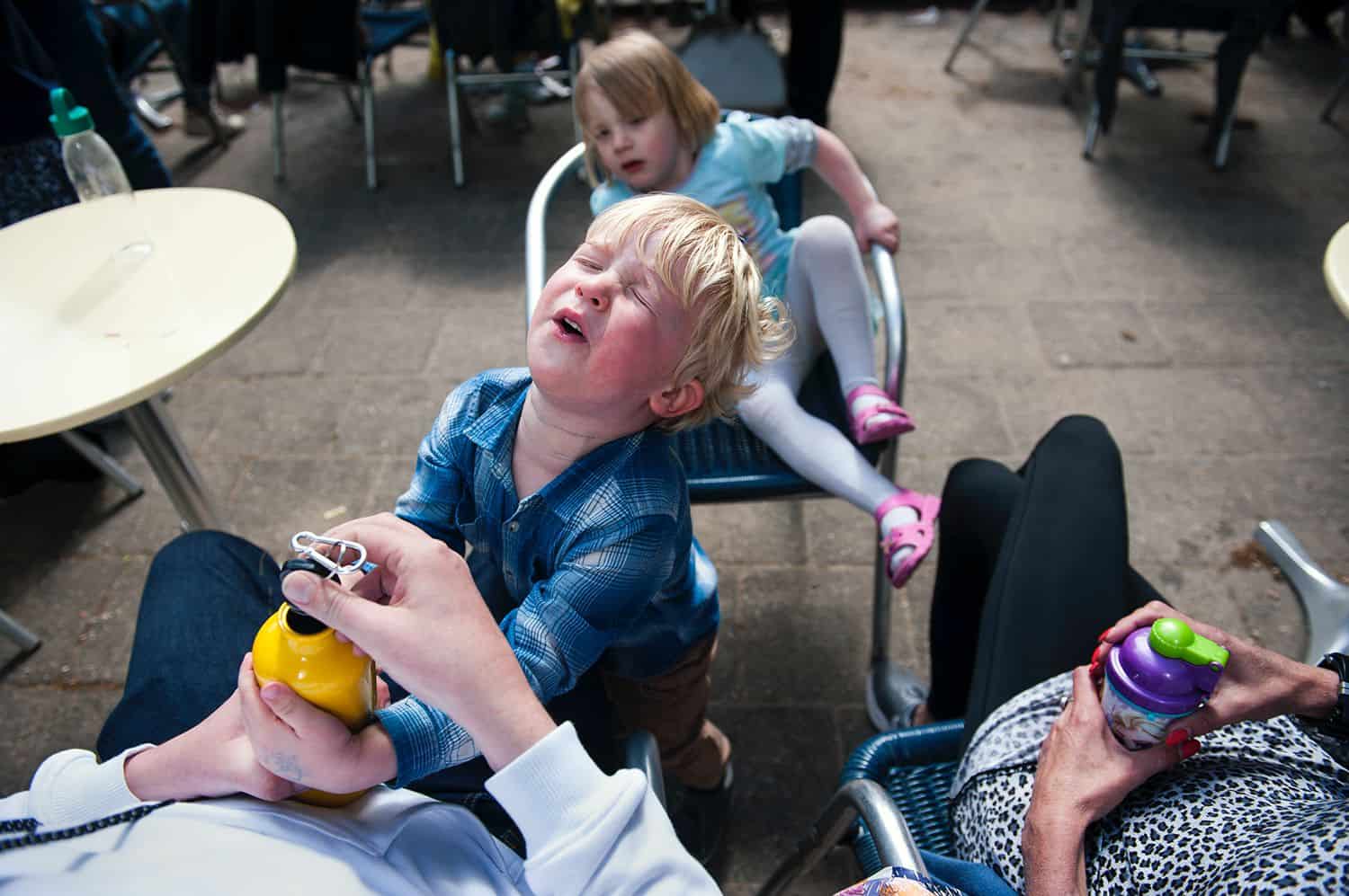 A little boy whines dramatically as his mother opens his yellow water bottle for him