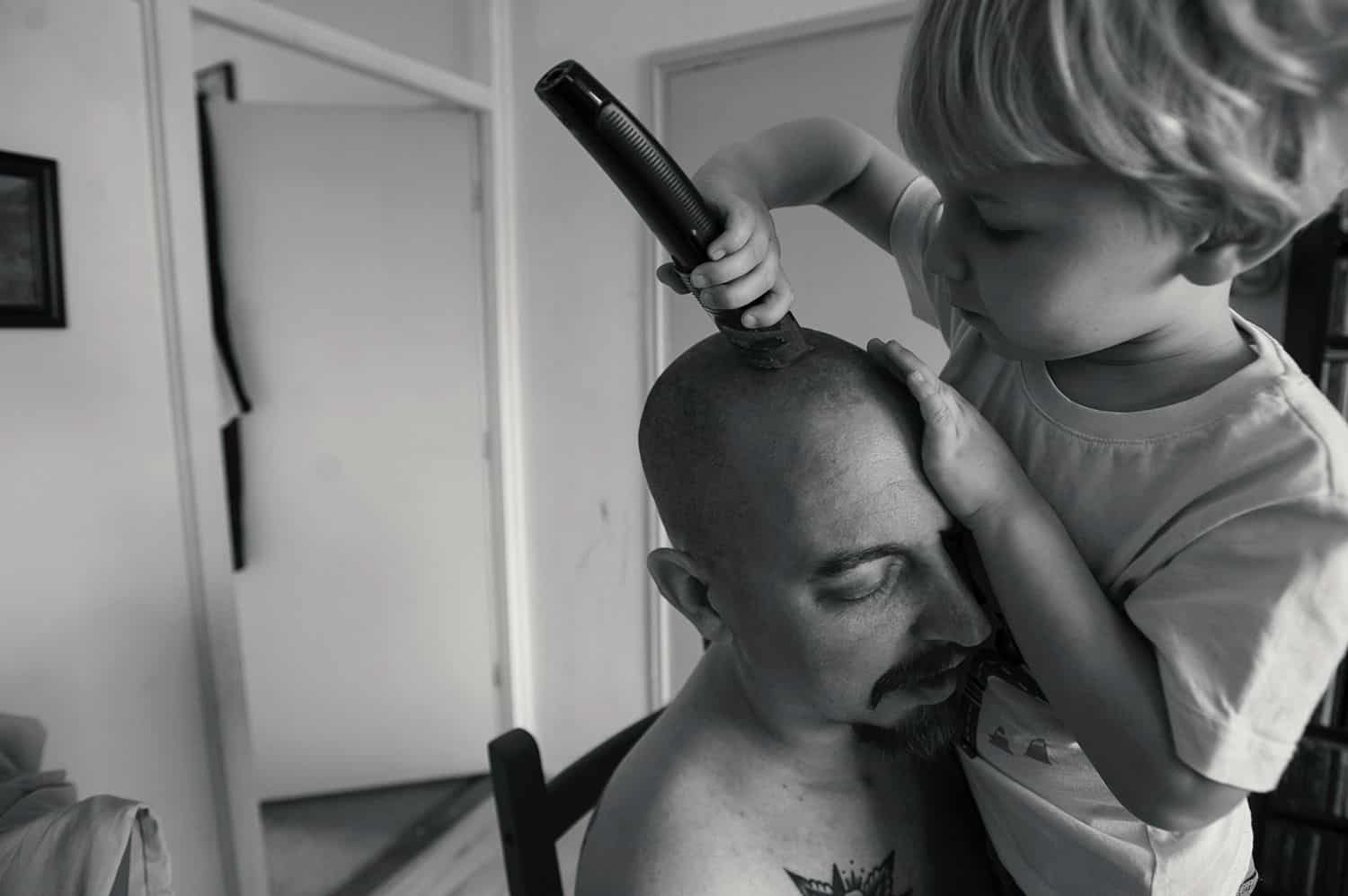 A dad holds his little boy close as the child shaves his father's dead with electric clippers