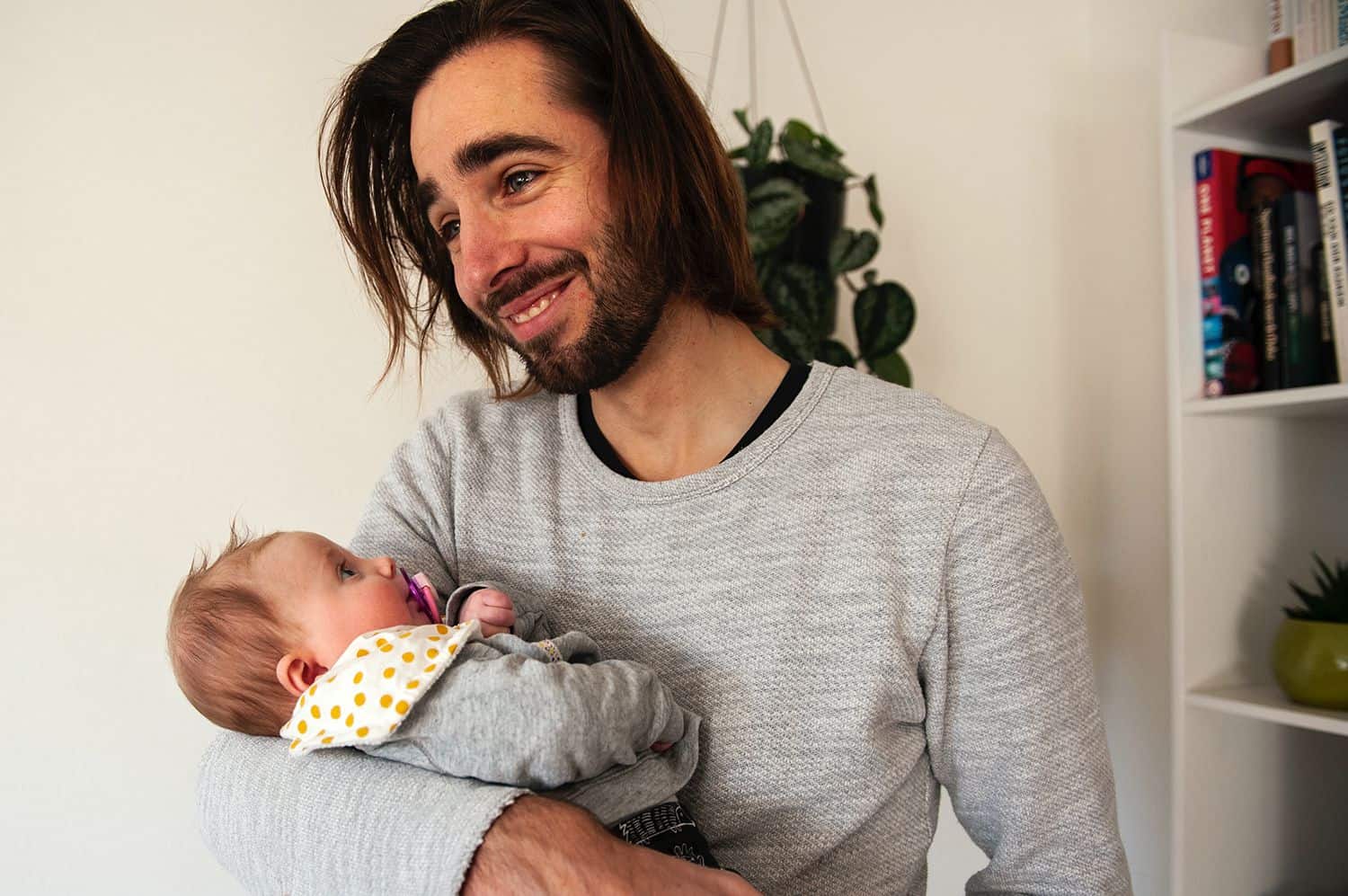 A dad smiles while holding his young baby who is sucking on a pacifier