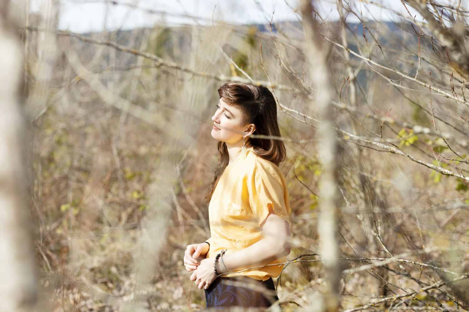 A person in a yellow shirt faces the sun while standing in the woods being photographed by Sofia Angelina