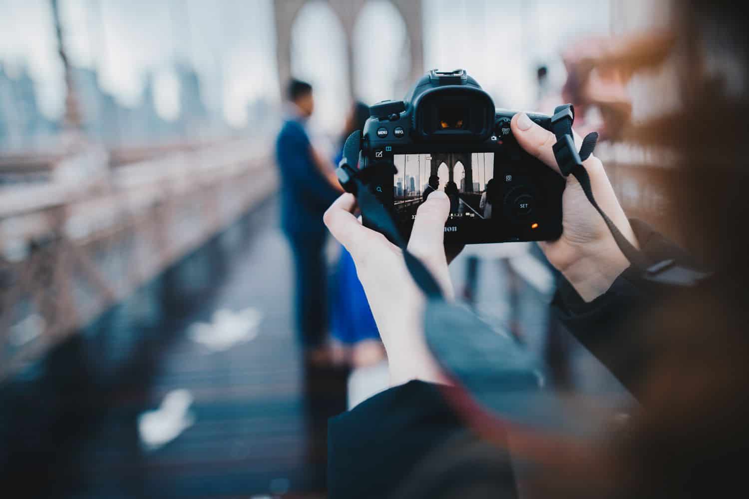 The back of a photographer's camera is in-focus as she photographs a couple standing on the Brooklyn Bridge, which is blurry in the background.