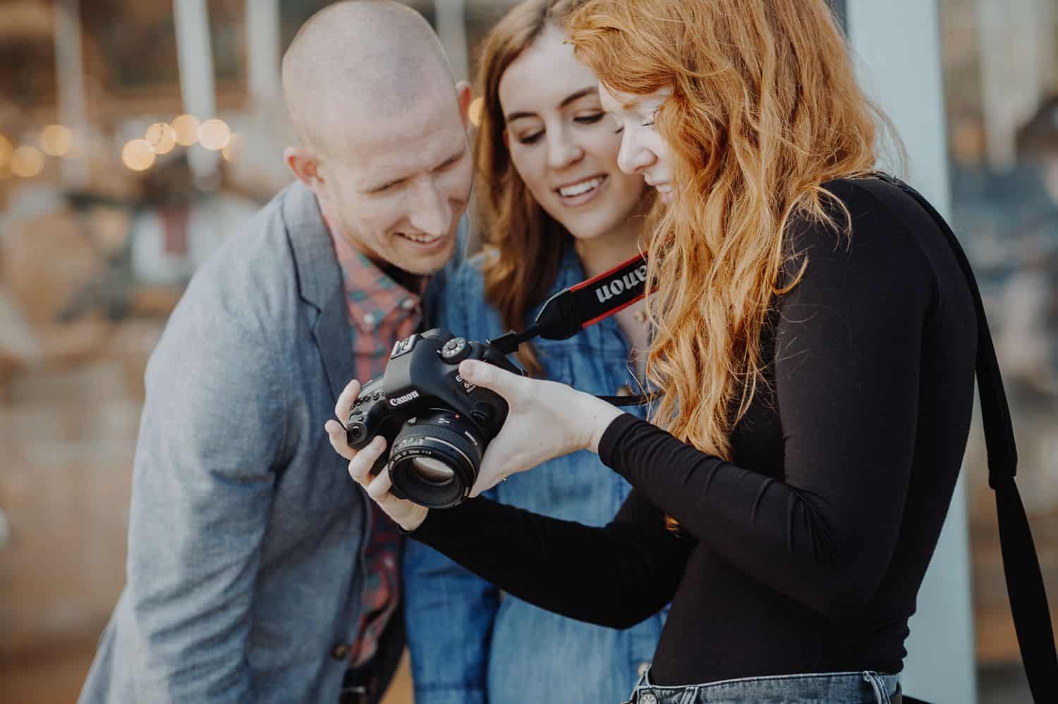 A photographer with red hair shows her camera's LCD screen to two happy clients.