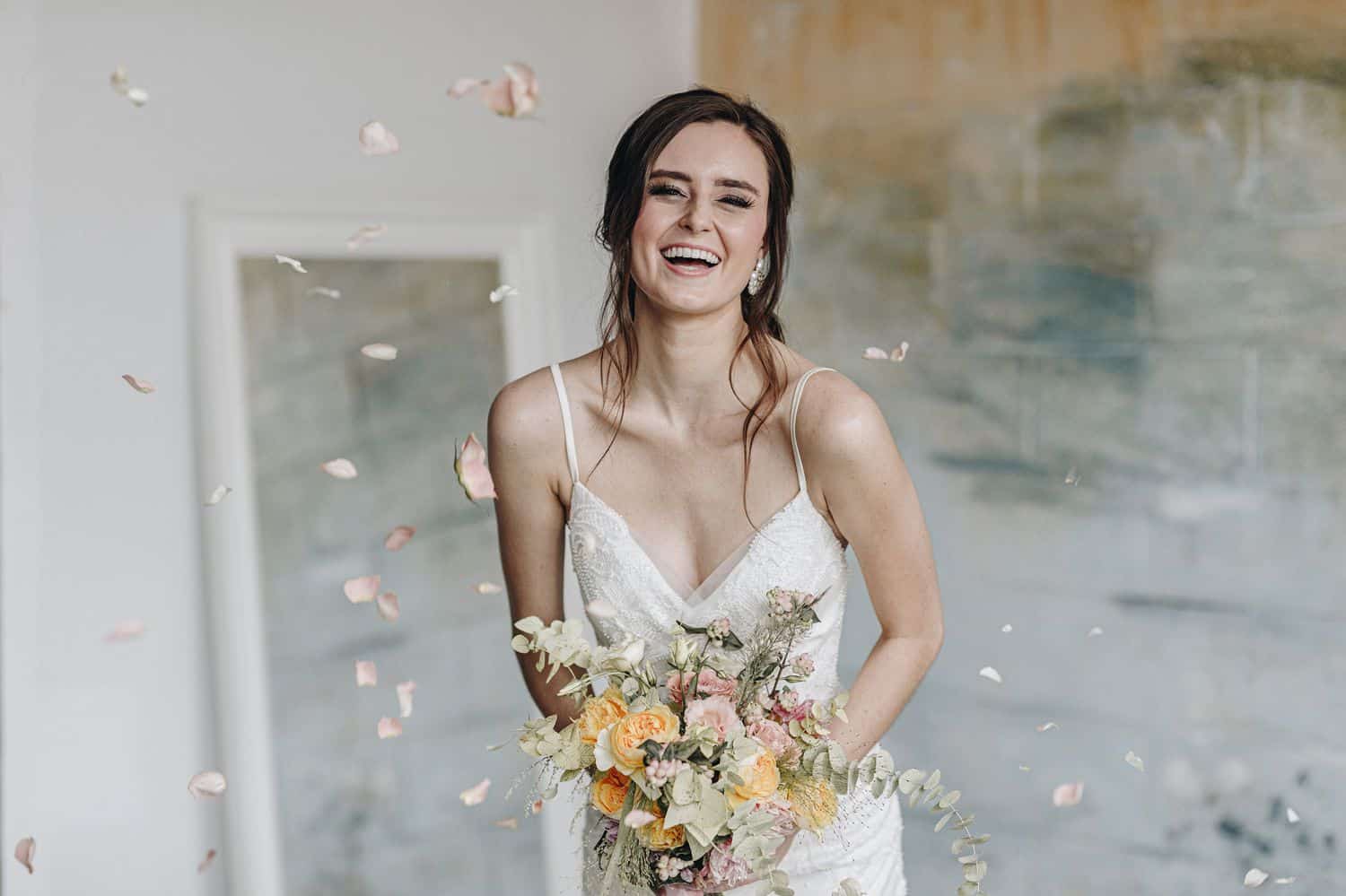 A brunette bride in a spaghetti strap wedding gown holds her bouquet and leans toward the camera laughing.