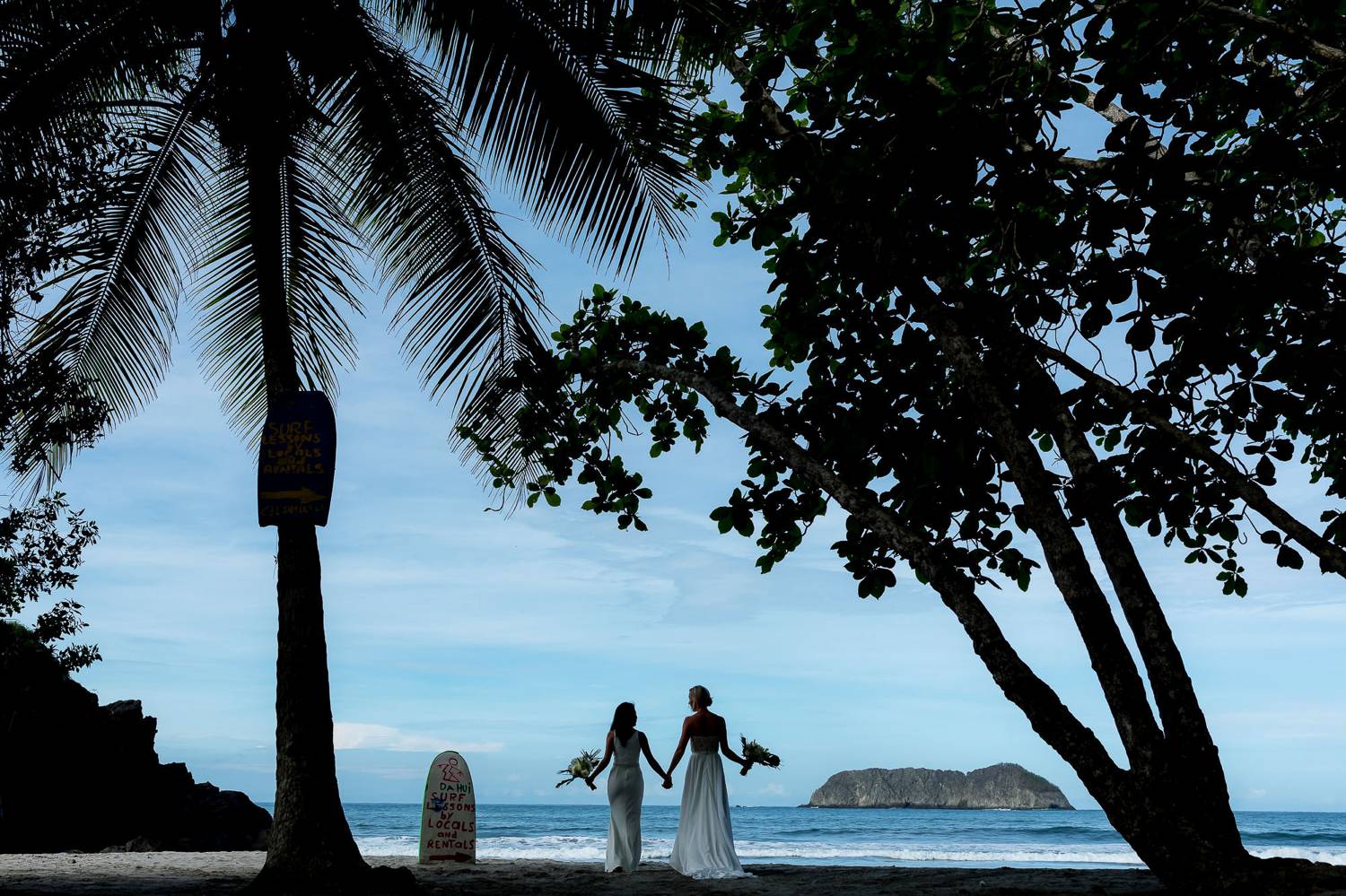 Client Experience: A photograph by Kevin Heslin depicts the sillhouettes of newlywed brides through palm trees as they walk along a beach at sunset.