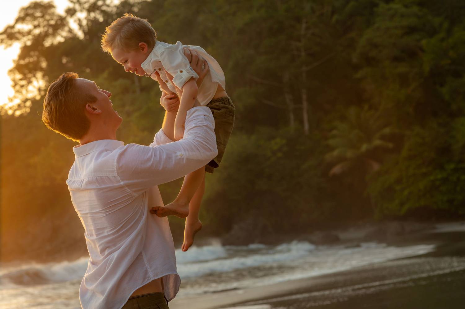 Client Experience: A photo by Kevin Heslin depicts a dad on a beach raising his child playfully toward the sky at sunset.