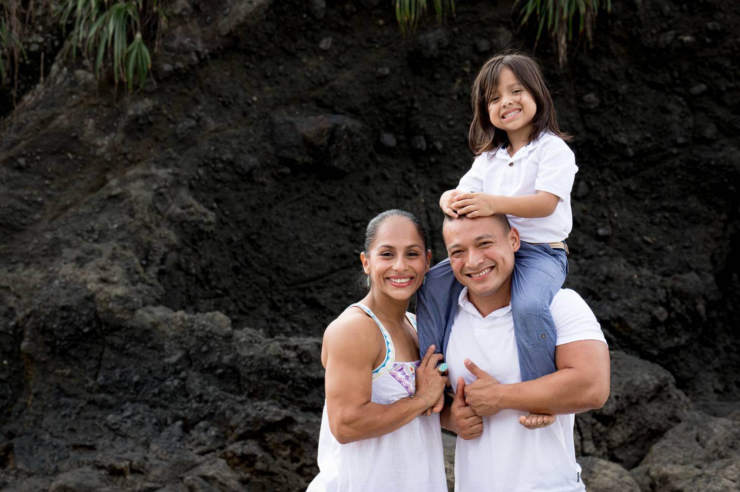 A photo by Kevin Heslin depicts two parents wearing white and posing with a smile as their child sits on the dad's shoulders.