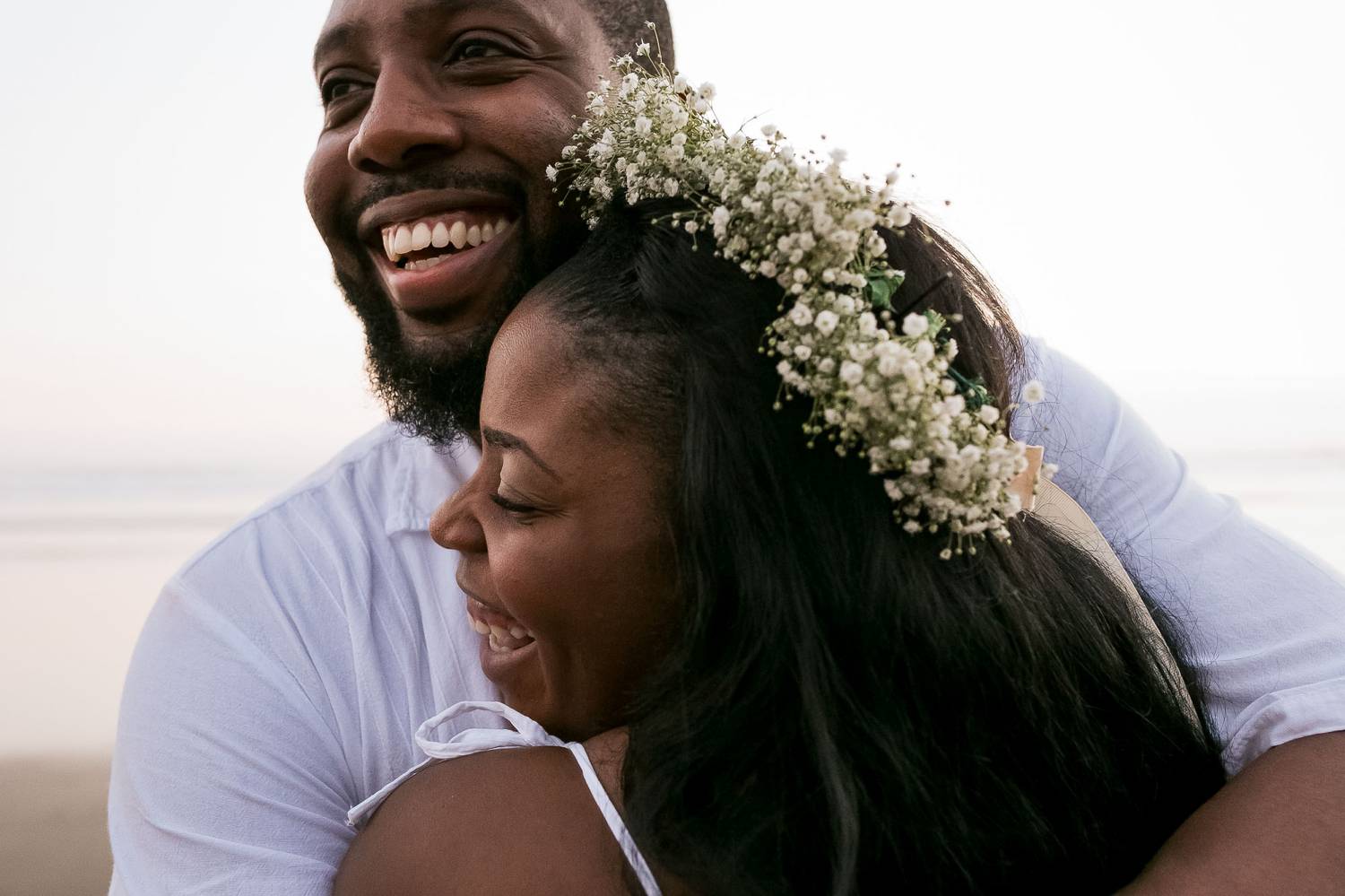 A close-up photo by Kevin Heslin depicts a Black bride and groom laughing and hugging on a beach.