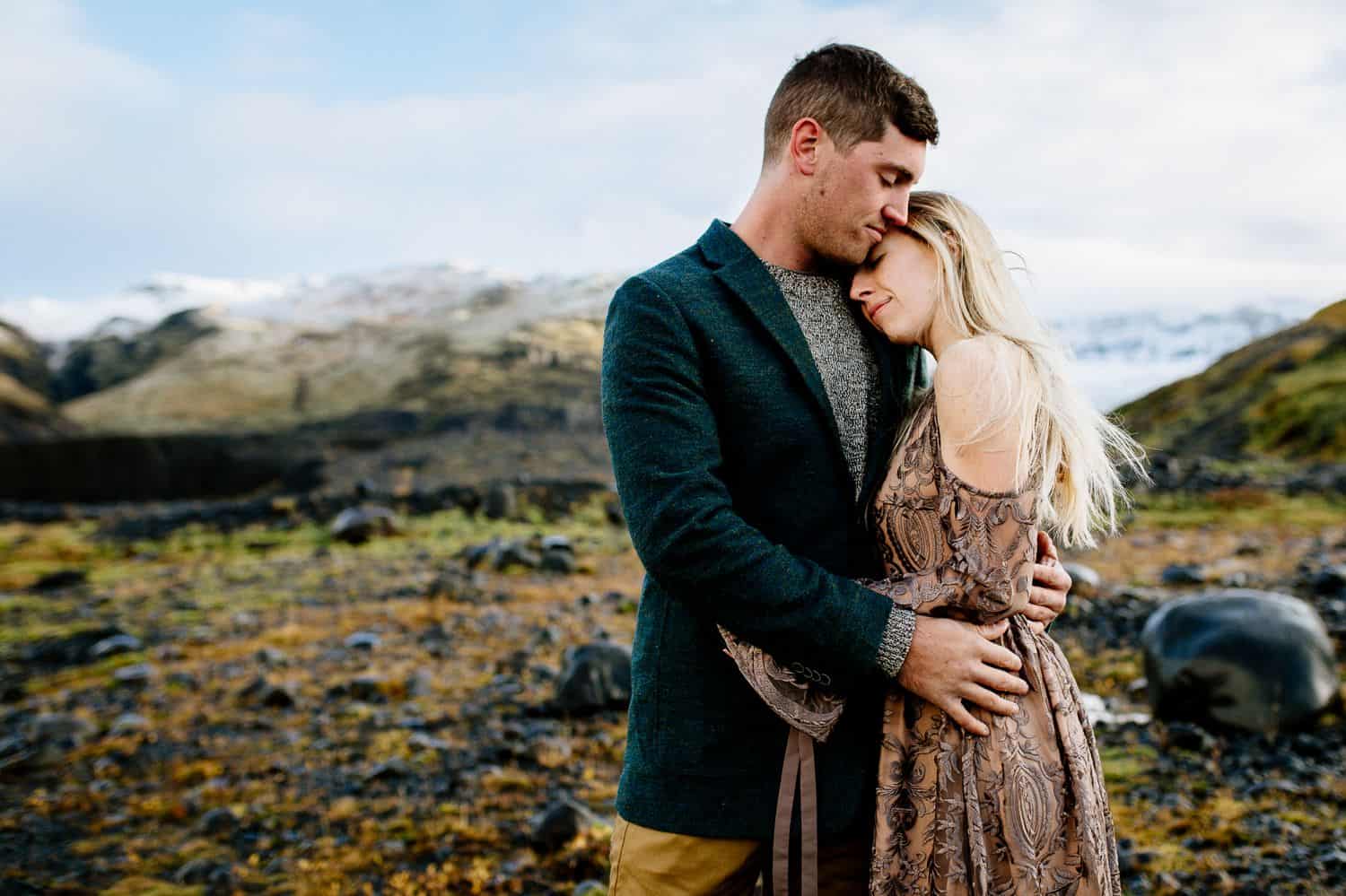 Man in a green jacket embraced a woman in a brown lace dress as they stand in the Icelandic landscape.