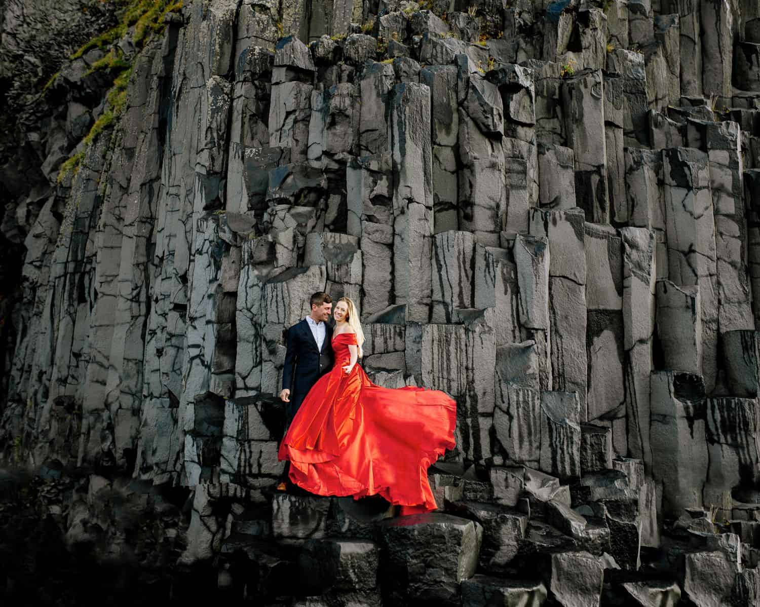 Man in a black suit and woman in a billowing red gown stand on a rock face in Iceland