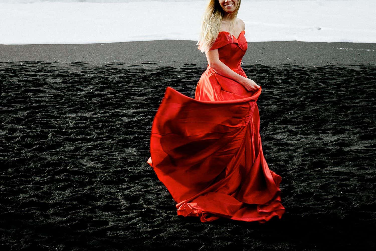 Woman in a flowing red dress dances on a black sand beach in Iceland. The photo is cropped so her eyes are not in the picture.