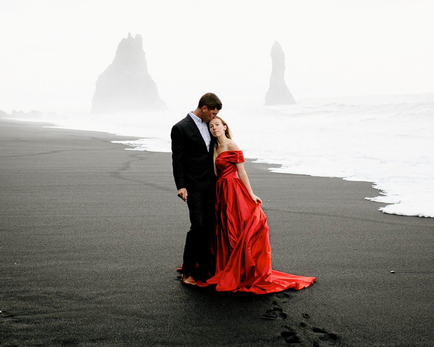 Man in a suit hugs a woman in a red dress on a black sand beach in iceland