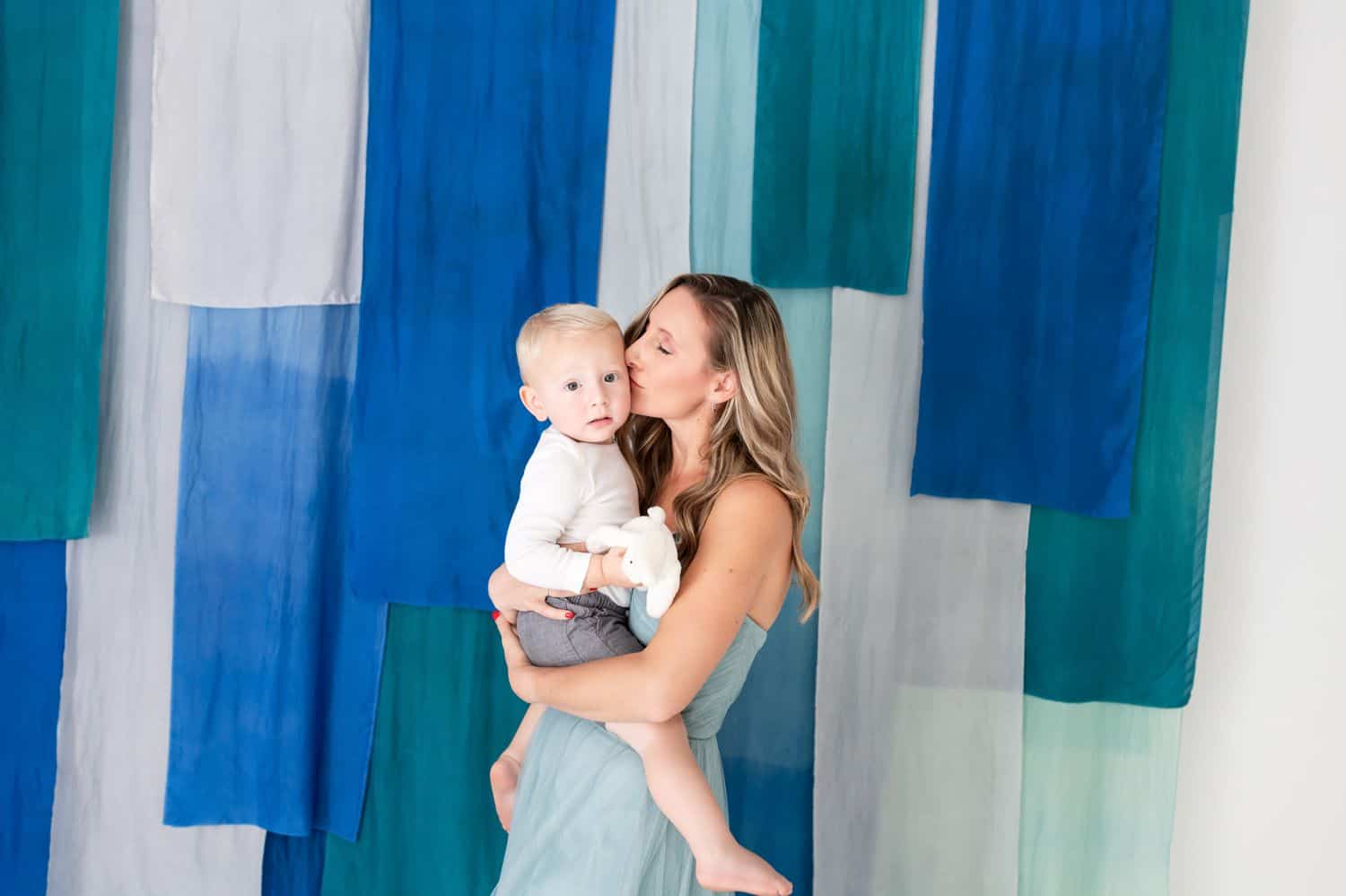 A mom kisses her toddler son on the cheek in front of a blue-draped wall.