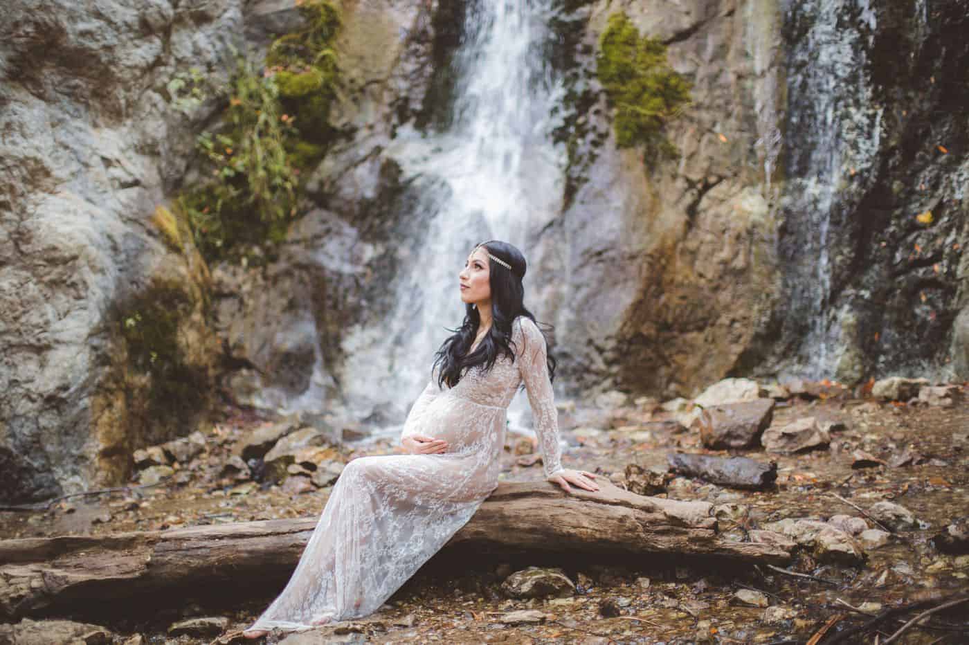 Vanessa Hicks photographs a pregnant mother sitting on a log in front of a waterfall.