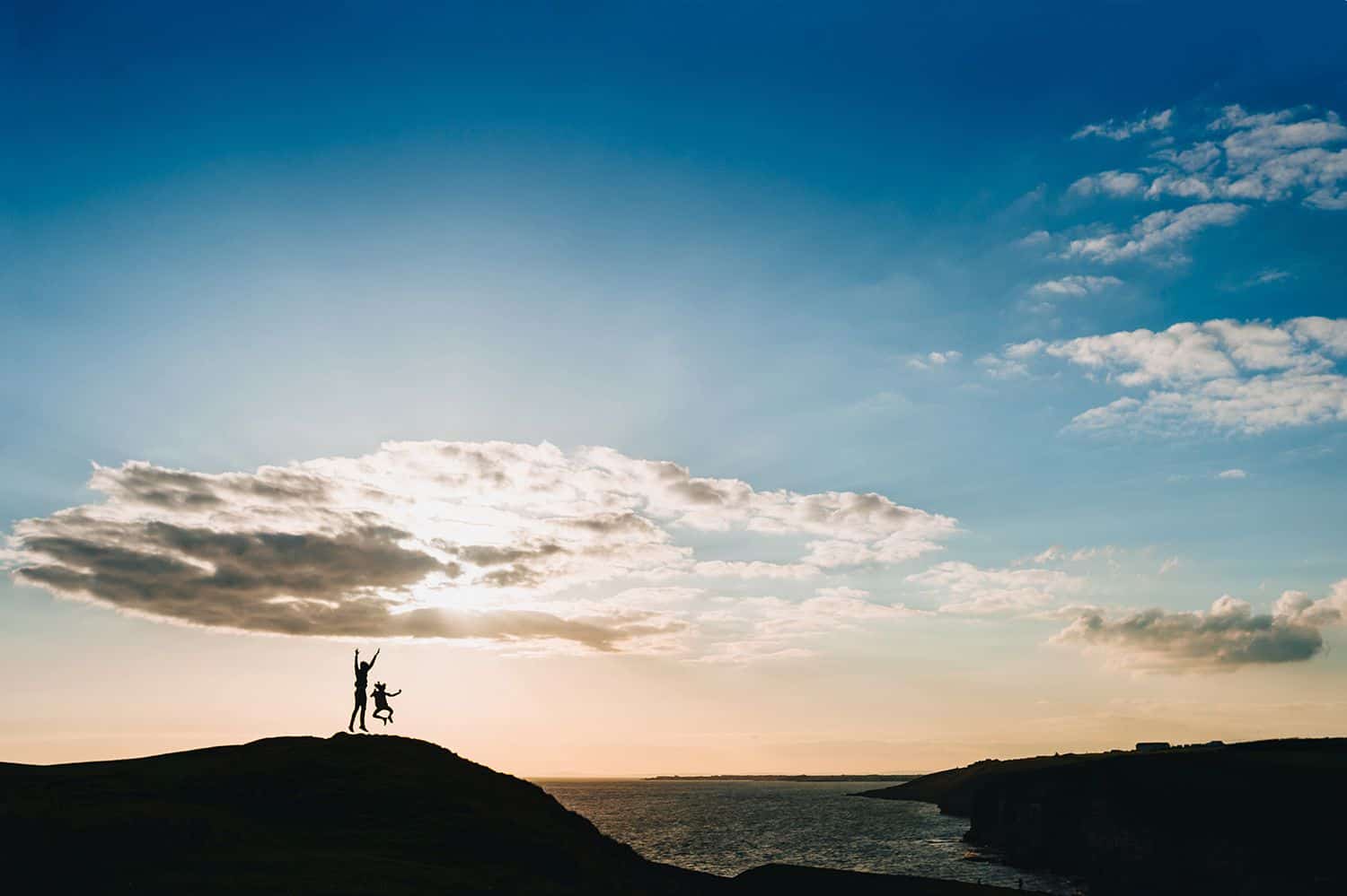 Silhouette of a parent and a child jumping on a hill overlooking the ocean.