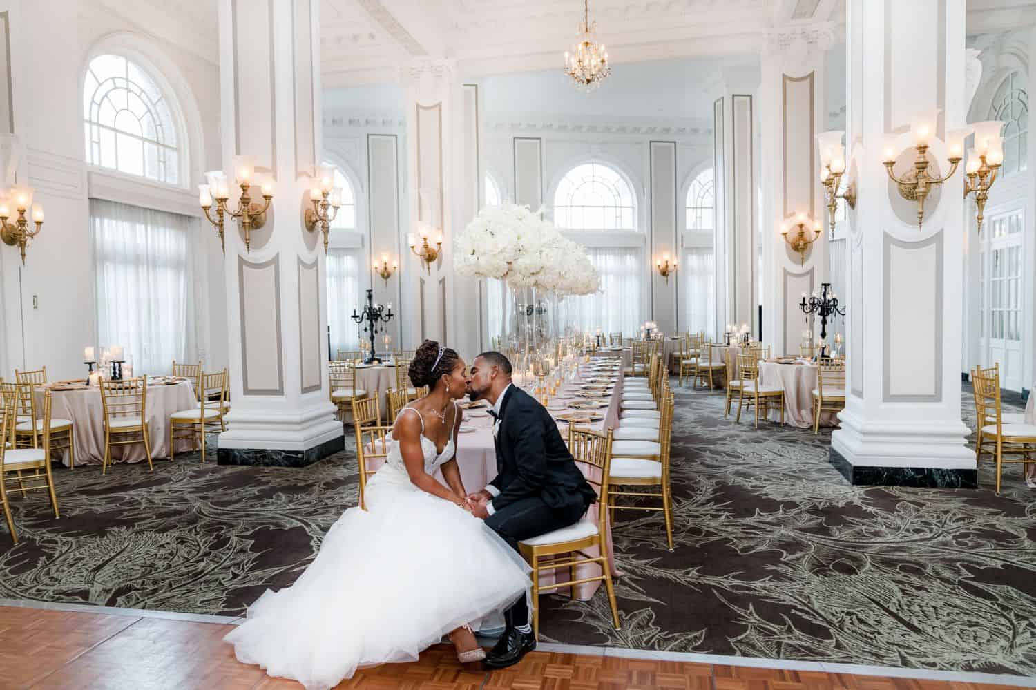 A bride in a white wedding gown kissed a groom in a black tuxedo. Both are seated in the front row of a ballroom full of chandeliers.