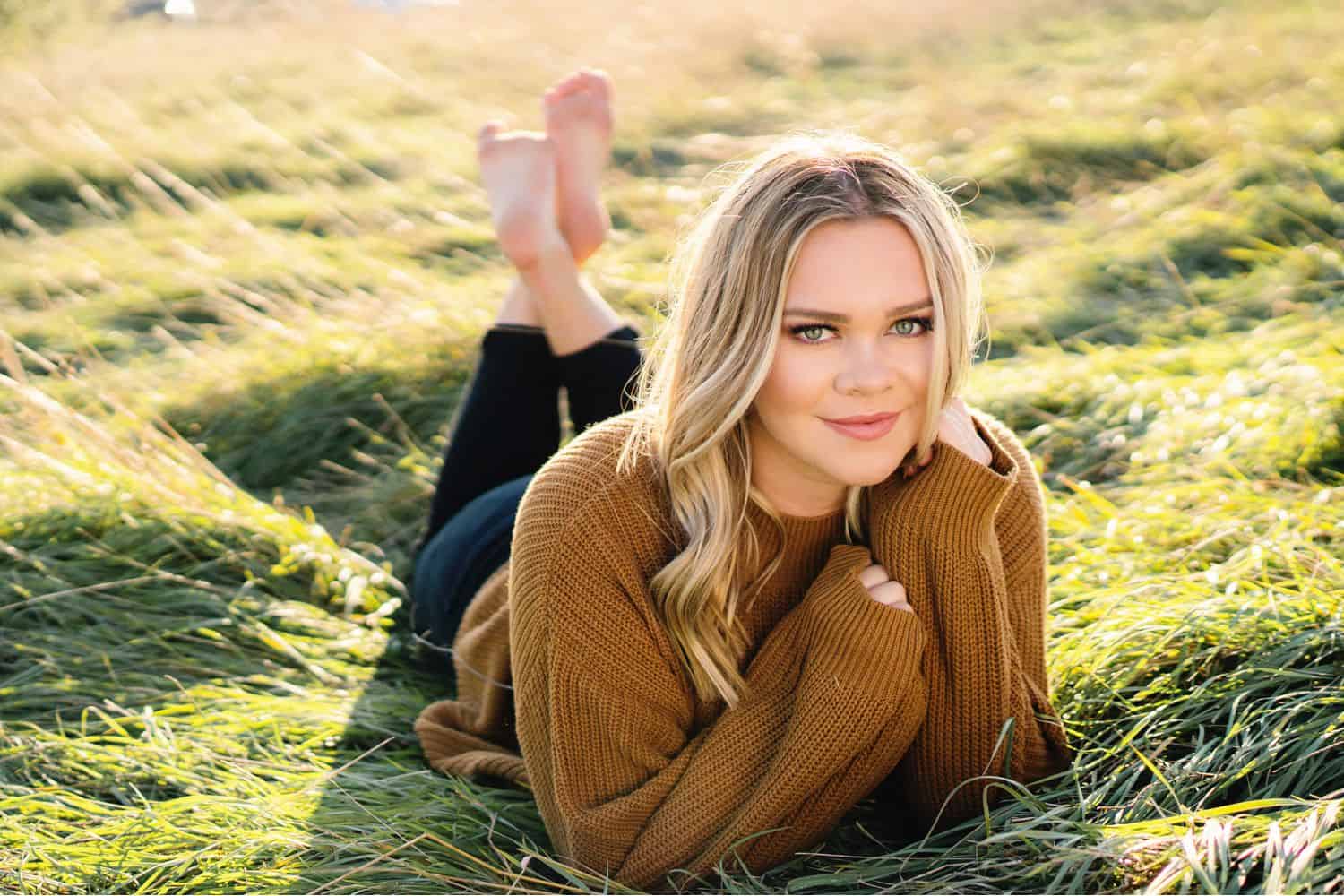 A senior girl in a gold sweater lies on her stomach in an early morning field with her chin in her hands