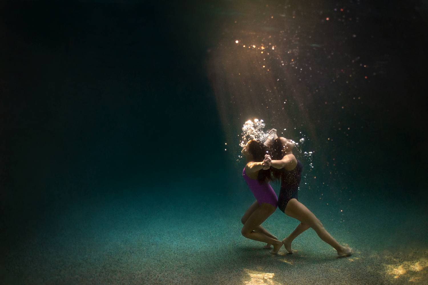 Two children stand back-to-back at the bottom of a swimming pool blowing air bubbles toward the surface