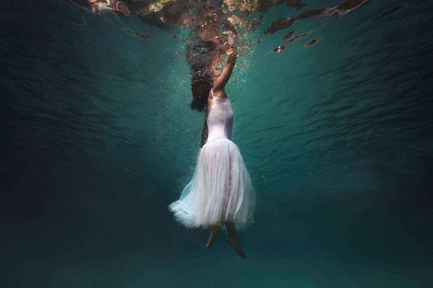 A person in a flowing white dress floats in a blue pool with only their face braking the surface of the water