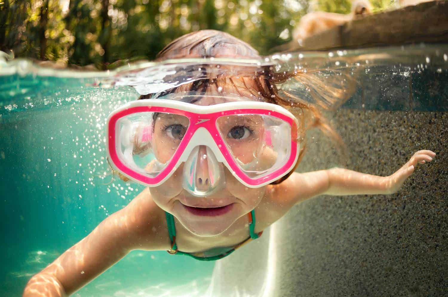 A child in oversized pink goggles swims underwater toward the camera