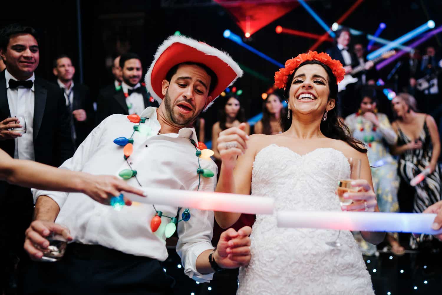 Wedding Print Packages: A bride and groom dance in silly hats at their reception. They are surrounded by guests holding glow sticks.